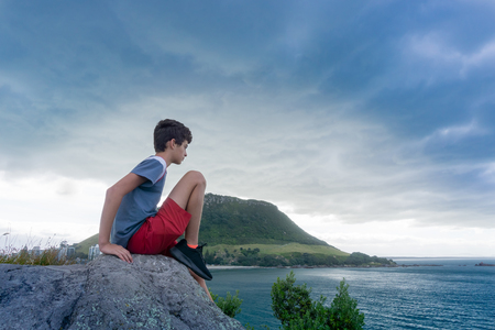Boy sits on rock on Leisure Island looking out to sea with Mount Maunganui  under evening light and dark clouds in backgroundの写真素材