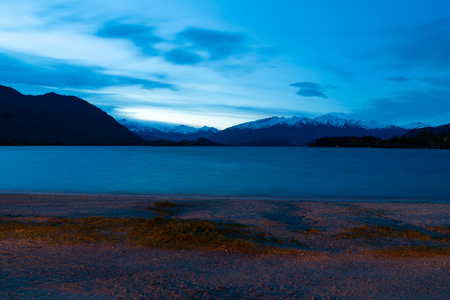 Nighttime view across Lake Wanaka to snow capped mountains under blue tone skyの写真素材