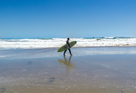MOUNT MAUNGANUI NEW ZEALAND - FEBRUARY 10 2019: At Main Beach male walks along in front of large dangerous surf carrying surf board boardのeditorial素材