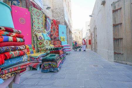 DOHA QATAR - JULY 11 2019; Brightly colored rugs with man in lane and traditional atire with...
