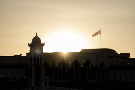 Lens flare and clock on tower as sun sets silhpoetting low buildings of traditional Arabic design.の写真素材