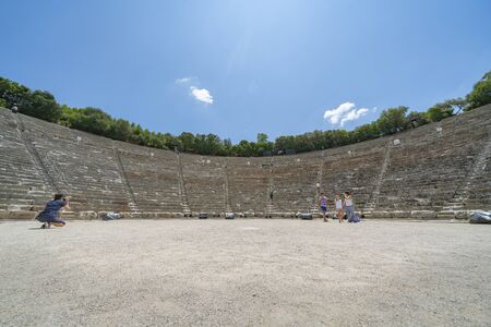 EPIDAURUS GREECE - JULY 19 2019; Tourists having photo taken on floor of Ancient Epidaurus theatre with semi-circular stone seating and numbered rows behind.のeditorial素材