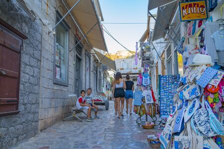 Hydra, Greek Island - July 20 2019; Three young women walking away while small boys sit on bench opposite souviners on display in narrow alleyway.のeditorial素材