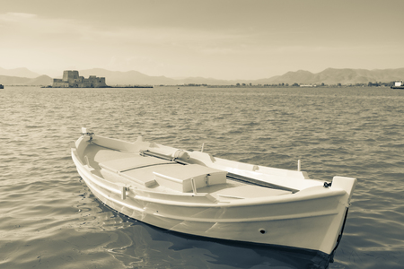 Old-fashioned sepia toned traditional style Mediterranean small boat afloat on sea at Nafplio with fortress on Bourtzi Island in background.のeditorial素材