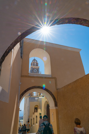 Santorini Greece - August 8 2019;  Sun blasts through under an arch in line up of architectural  features of arches, bell tower and clock at Fira, as tourists walk through.のeditorial素材