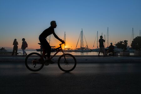 Naxos Greece - August 11 2019;  in silhouette a boy cycles past in foregropund while tourists look at view and locals fish in morning on Naxos town waterfrontのeditorial素材