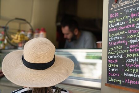 Chalki Naxos - August 13 2019; Wide brimmed hat of woman waiting to order and receive their icecream through a hole-in-wall shop on warm Greek Island day.のeditorial素材