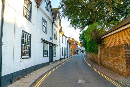 Shady narrow street with white and black buildings on one side and red brick wall on other side and yellow  lines leading through town.の写真素材