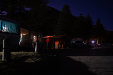Row of holiday-making campers light through caravans an tents during night along beachfront sits...