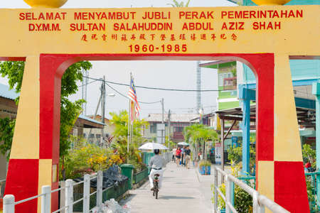 Palu Ketam Malaysia - October 10 2013; Bright red and yellow welcome sign at Crab Island floating fishing village off Malayasian coast people on walkway between piers and buildings through archのeditorial素材