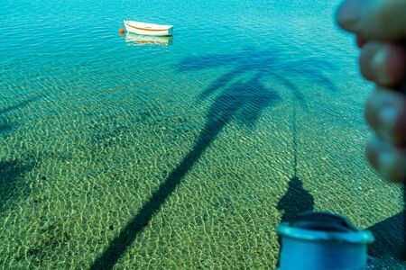 Idyllic coastal scene of calm shallow water with long diagonal palm tree shadow and small boat floating beyond the palm fronds with persons holding a fishing rod casts shadow on turquoise water.の写真素材