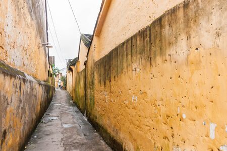 Long alleyway leading to open area in distance  between buildings with walls weathered and growing green algae in city Hoi An, Vietnamの写真素材