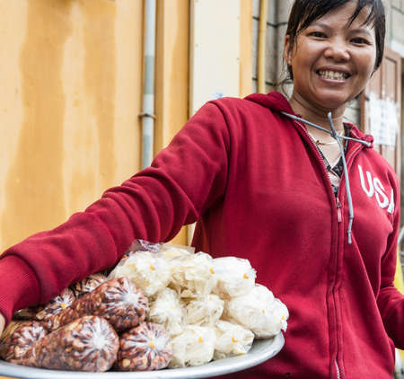 Hoi An Vietnam October 17 2020; Woman street vendor smiling and holding tray of bagged peanuts and popcorn for sale in city streetのeditorial素材