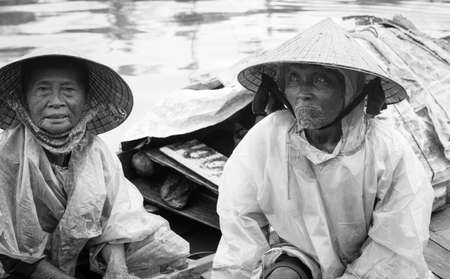Hoi An Vietnam October 17 2020; Senior Vietnamese couple sitting their small boat in river sheltering from rain in conical hats and plastic covers in monochrome.のeditorial素材
