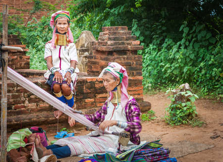 Bagan Myanmar - October 29 2013; Two Kayan women with brass rings around necks one weaving while other in selective focus sitting wall looks on.のeditorial素材