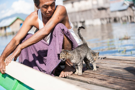 Inle Lake Myanmar - November 2 2013; Man crouching on pier holding edge of boat with two cats.のeditorial素材