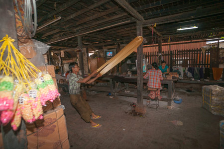 Mandalay Myanmar - November 5 2013; Confectionary factory with man pulling and stretching long string of toffee while women workers pack on bench in background.のeditorial素材