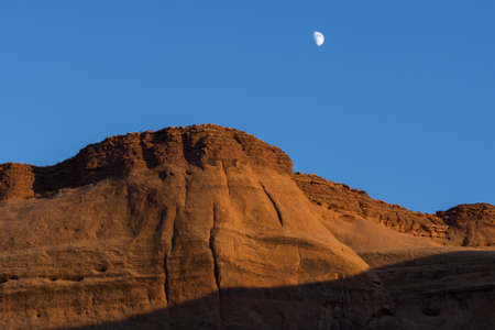 Sun lowers in clear blue sky and brightens a red rock escarpment under white moonの写真素材