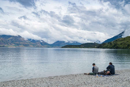 Queenstown New Zealand - February 28 2015; Three young adult travellers relaxing in early morning light on shore of Lake Wakatipu in low light split toned effect.のeditorial素材