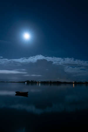 Darkness of the night long exposure in moonlight with white clouds drifting across dark blue sky over still of bay below.の写真素材