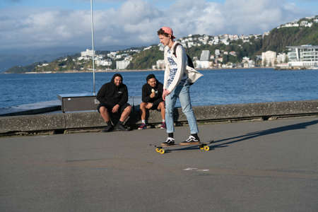 Wellington New Zealand - July 29 2021; Late afternoon long shadows and people passing on old wharf promenade on city waterfront with couple sitting on edge as skateboarder with cap on backwards skates by.のeditorial素材