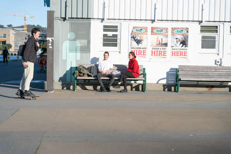 Wellington New Zealand - July 29 2021; People seated talking as young man skateboards past old wharf shed with leisure equipment hire advertising posted on wall on city waterfront areaのeditorial素材