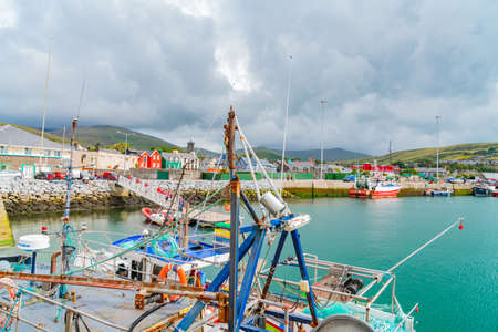 Dingle Ireland - August 12 2017; Commercial fishing boats moored on one side or bay with homes businesses and rolling green farmland across on other side.のeditorial素材