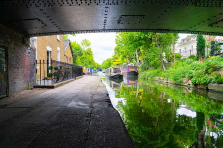 London - England - August 5 2017; From under a bridge looking into the day narrow boats and canal activity along London's Regent's Canal. and reflections from trees along edge and homesのeditorial素材