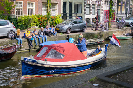Amsterdam-Netherlands - August 21 2017; Quaint small blue boat with red canopy carrying Dutch flag cruising on canal past group young adults sitting on wallのeditorial素材