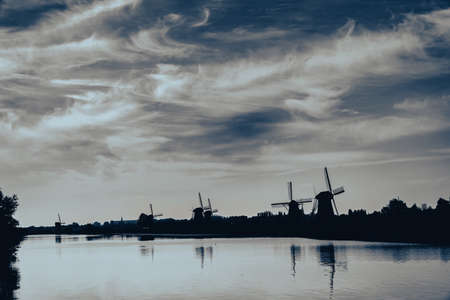 Row silhouette windmills along waterway under changing cloudy sky in Kinderdijk, Netherlands.の写真素材