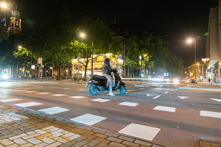 Rotterdam Netherlands - August 23 2017; Young woman on motor-scooter crosses cautiously under night street lightsのeditorial素材