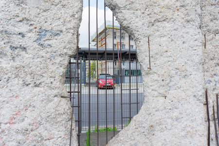 Berlin, Germany - September 25, 2017: Scene through Berlin Wall hole broken with reinforcing iron view out onto street with red car entering street.のeditorial素材
