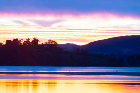 Land and hills in silhouette across bay back-lit at sunrise with dramatic bright sky colors reflected in calm water of Tauranga Harbour.の写真素材