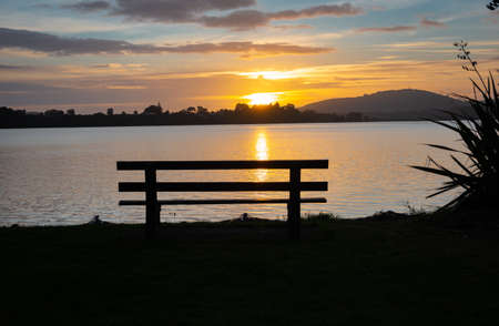Tauranga sunrise, across bay from empty bench seat in silhouetteの写真素材