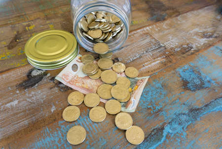 Savings jar with New Zealand currency coins and bank note spilling out on to wooden table closeup.の写真素材