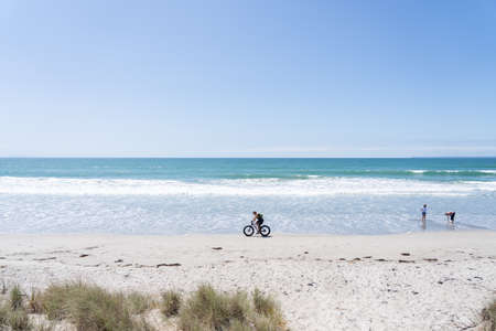 Papamoa New Zealand - February 22 2022; Cyclist near water's edge rides past two women on beach.のeditorial素材