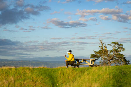 Tauranga New Zealand - February 25 2022; Woman in bright yellow top resting on walk around the Mount taking photos on mobile phoneのeditorial素材
