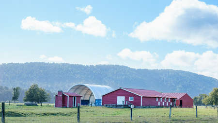 Red shearing shad and small building on Westland farm with hills backdrop under cloudy sky.のeditorial素材