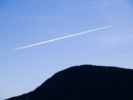 White jet stream through clear blue sky over back-lit silhouette hill