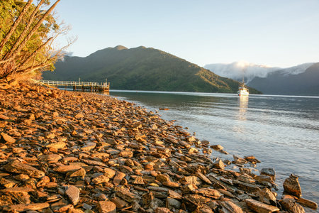 Stony foreshore in Marlborough Sounds bay at sunrise with launch at anchor in middle.の写真素材