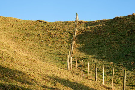 Fence leading up and over slope under blue sky in agricultural background.