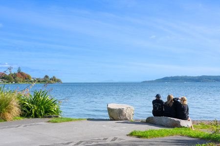 Taupo New Zealand - April 16 2023; Three young adults sitting by lake looking at view.のeditorial素材