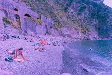 Riomaggiore Italy April 26 2011; Stony beach with sunbathers below stone wall by sea in blue hue...