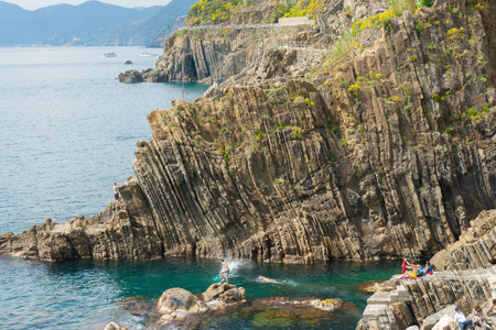 Riomaggiore Italy April 26 2011; Sunbathers watch youth jumping and diving from high rock cliff sea...