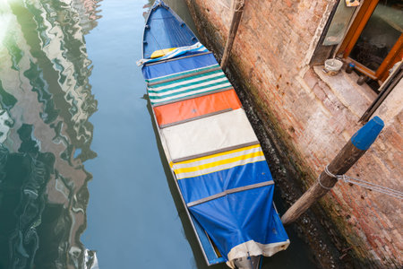 Colourful cover on boat tied-up below in canal beside rustic brick wall and window.の写真素材
