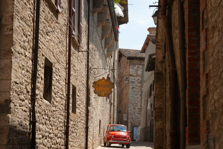 Gubbio Italy - May 13 2011; Narrow shady street lined by tall European buildings with red small car under sign for tavernのeditorial素材