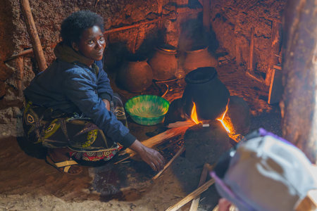 Gasura Rwanda - September 8 2025; Authentic African village life woman cooking over open fire squatting on floor of small dark hut cooking on open fire looking up.の写真素材