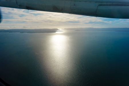 Long sun-trail across sea from distant coastline under wing of plane while in flight around New...