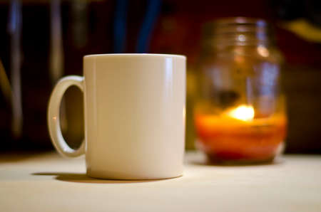 A single blank white coffee mug on a wooden work bench under the light of a candle getting ready for the morning work.の写真素材