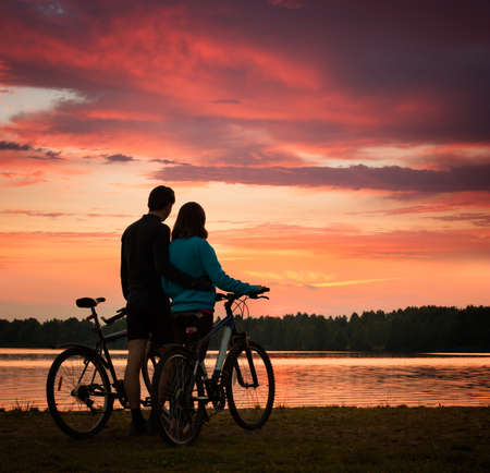 Romantic Couple with Bicycles Watching Sunset. Summer Nature Background with Beautiful Clouds, Sky and River. Active Leisure Concept.の写真素材